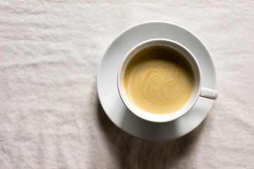 High Angle Still Life of Frothy Coffee in White Cup with Saucer on Table Covered with White Tablecloth and Copy Space