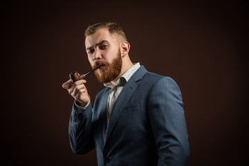 Portrait of handsome confident man in suit holding smoking pipe against of brown...