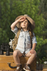 Adorable little girl in a safari hat and explorer clothes playing safari sitting on wooden suitcase outdoor. Children's play concept. Looking for the summer vacation