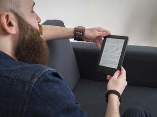 Young bearded man reading e book on the sofa. Shot from behind.