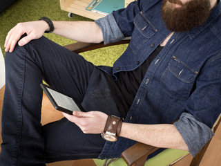 Young bearded man reading e-book at home sitting on a chair. No