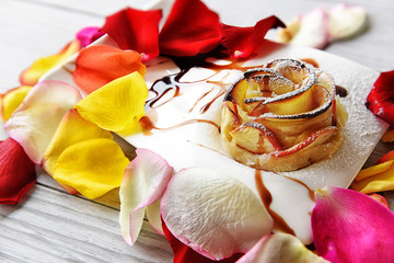 dessert. apple baked in the shape of a rose. Around colored petals of roses. Wooden light natural background
