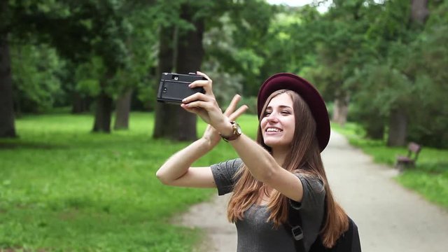 Girl Tourist Makes Selfie on Old Film Camera. the Woman is Very Beautiful. She is Wearing a Red Hat and a Gray Dress. in Terms of Many Green Trees.