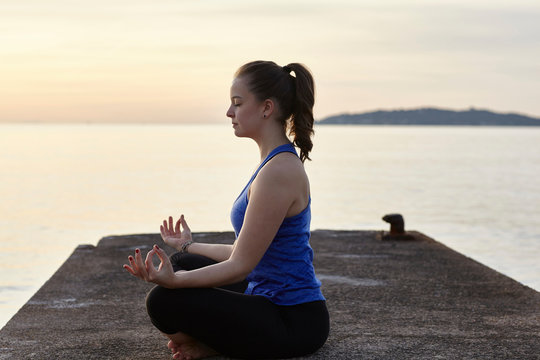 Young woman sitting on pier, in yoga position, at sunset