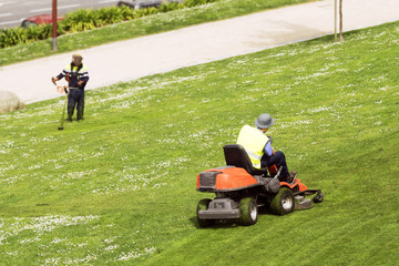 utility workers of municipality  with  brush cutting  and   lawn  mower , riding  mower,  in city park ; selective focus