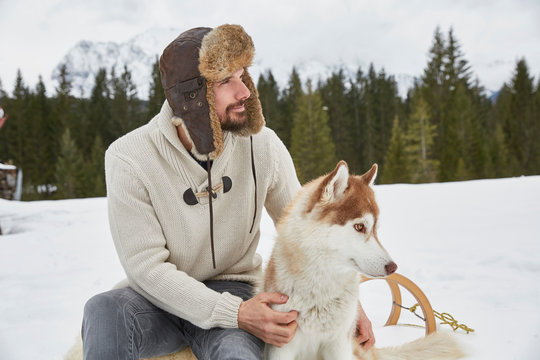 Young man wearing trapper hat petting husky in snow, Elmau, Bavaria, Germany