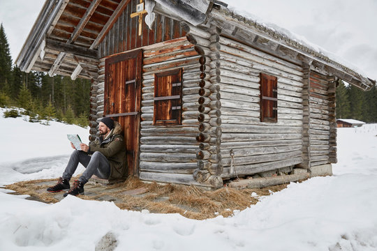 Young man reading map sitting outside log cabin in winter, Elmau, Bavaria, Germany