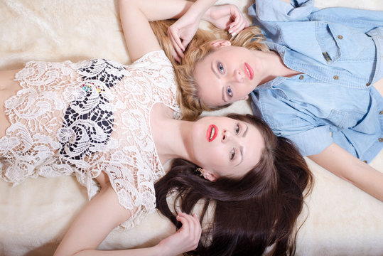 picture of 2 glamor blond & brunette girlfriends with red lips having fun posing lying in bed together head to head in blouses & looking at camera relaxing on light copy space background