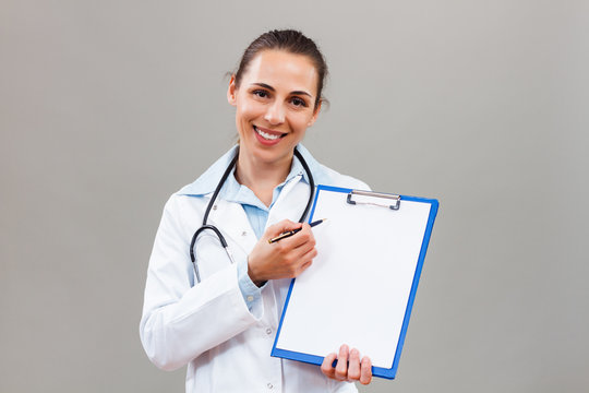 Beautiful female doctor is holding clipboard  and showing document with pen.