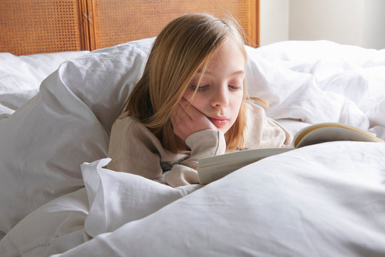 Blond haired girl reading book in bed