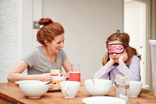 Mid adult woman at breakfast table with daughter wearing sleep mask