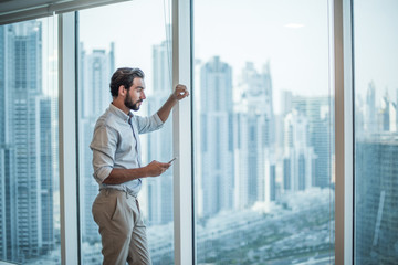 Businessman with smartphone staring through window with skyscraper  view, Dubai, United Arab Emirates