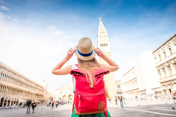Young female traveler with hat and backpack standing on San Marco square with tower and basilica on the background in Venice. Back view - obrazy, fototapety, plakaty