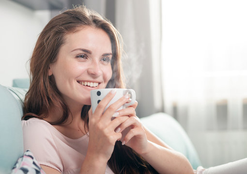 Smiling woman sitting on couch at home and drinking coffee. Casual style indoor shoot
