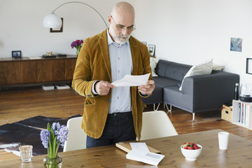 Mature man standing by table and reading letter at home
