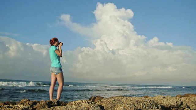 Young woman with red hair in glasses photographs sea standing on the rocks in front of clouds , beach of Dominican Republic, wide angle