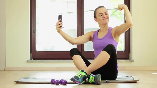 Portrait of cheerful woman using mobile phone to take selfie picture while showing her biceps at gym