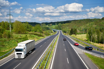 Asphalt highway with a moving white truck between lush forests in the countryside. Passenger cars entering the highway. In the distance the bridge and Electronic toll gate. View from above.