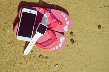 Flipflops, mobile phone and smart watch on sandy ocean beach vacation of concept, top view flat lay style
