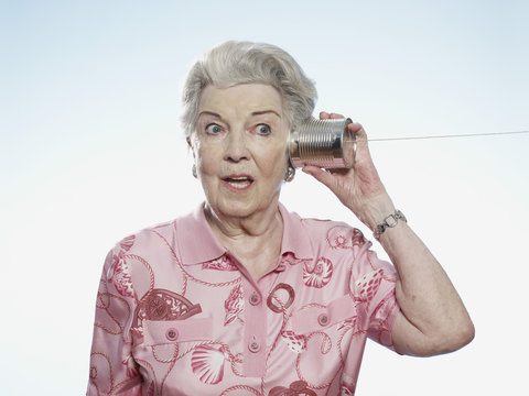 Senior woman comically strains to listen as she holds a tin can phone to her head