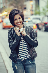 Portrait of beautiful Hispanic latin girl woman short black hair in leather jacket with headphones outside in evening night city street smiling laughing looking in camera, lifestyle portrait concept