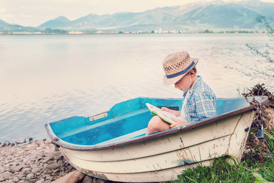 Boy with book seats in old boat on the lake bank
