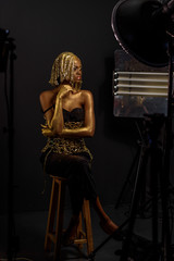 Independent black woman wearing gold dress, headdress and evening make up with lots of shine sitting on wooden chair in dark studio