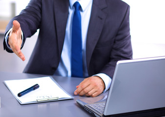 businessman sitting at desk holds out his hand for a handshake