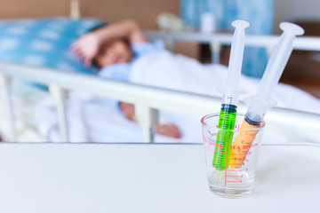 Syringes in a glass measuring cup with blurred illness boy lying