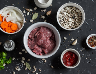 Raw ingredients - beef, beans, onions, carrots, spices and herbs, tomato sauce on a dark stone background