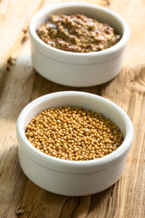 Yellow mustard seeds and whole grain mustard in small bowls, photographed on wood with natural light (Selective Focus, Focus one third into the seeds)