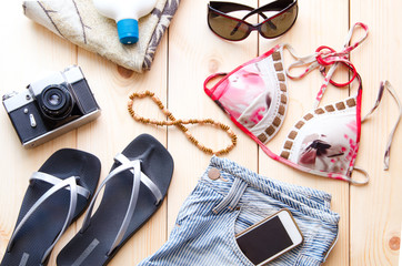 Summer accessories for travel or vacation concept. Vintage camera, denim shorts glasses, bikini, flip flop, beach towel,  sunburn lotion,  mobile on wooden background.