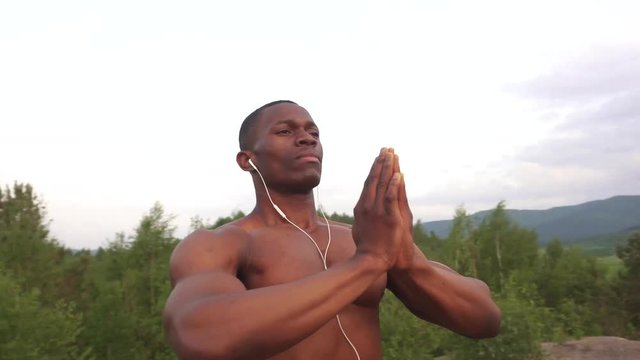 Muscular african american body builder with prayer hands stretching on the mountain peak during his morning outdoor training. Sunrise sky background. Moment of harmony and happiness