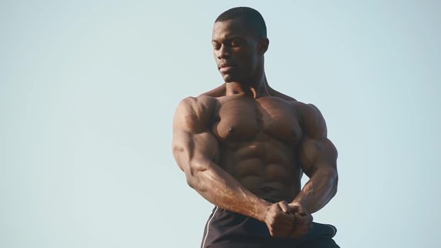 Strong handsome african american bodybuilder showing his perfect muscles on the clear blue sky background