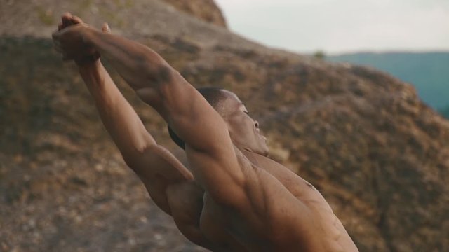 Close-up of african american bodybuilder with naked torso stretching outdoor. Rocky mountain landscape background
