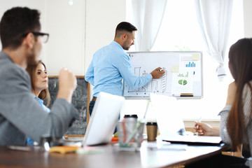 Obraz premium Handsome young man in glasses standing near whiteboard and pointing on the chart while his coworkers listening and sitting at the table