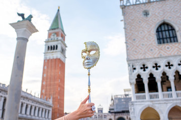 Fototapeta na wymiar Female hand holding Venetian carnival mask with San Marco tower on the background