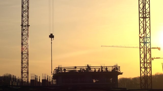 Silhouettes of cranes and construction site workers against orange sunset. 4K