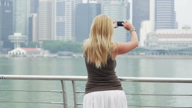 young woman having fun takes pictures and doing self portrait with smartphone  smiling at the camera with modern city skyscrapers in the background at singapore bay