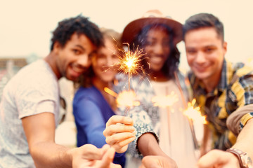Fototapeta premium Multi-ethnic millenial group of friendsfolding sparklers on rooftop terrasse at sunset