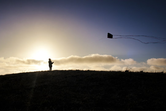 Child (10-11) flying kite on hill at sunset