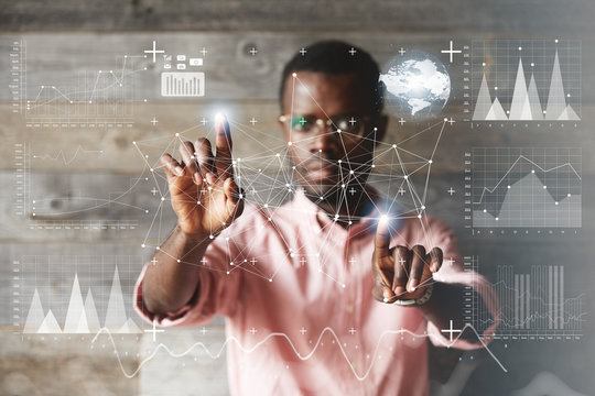 Double exposure. Black young businessman wearing glasses and casual shirt, touching futuristic screen interface with his fingers, making calculations, analyzing diagrams and schemes. Selective focus