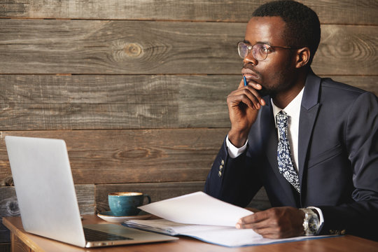 Handsome Young African Man Wearing Formal Suit Sitting At A Coffee Shop With Pensive Look, Thinking Of Business Plans, Holding A Pen, Leaning His Elbow On The Table, Signing Papers, Working On Laptop