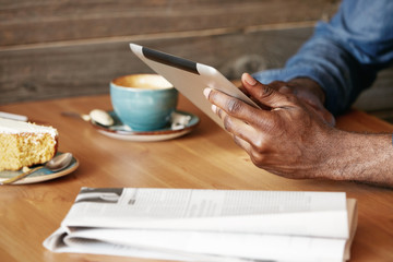 Close up view of African man's hands holding digital tablet, typing a message via social networks. Young black freelancer using electronic device for distance work while sitting at a cozy cafe.