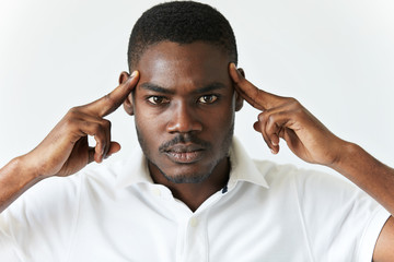 Black man in white polo shirt having bad headache. Young African American male thinking over or trying to recollect something very important, holding hands on his temples, looking at the camera.