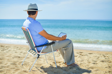 Man using tablet relaxing on ocean beach, blue sky outdoors background