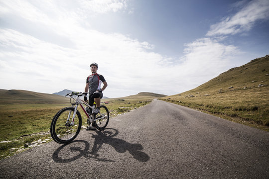 Young man posing with bike landscape with hills and asphalt road in diminishing perspective