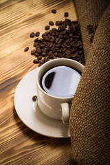 Coffee beans and coffee in white cup on wooden table with burlap