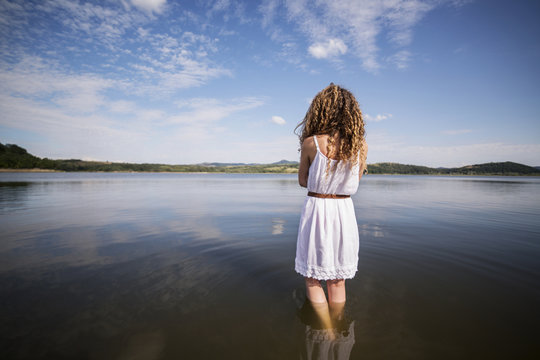 Rear view of female with curly hair, wearing white dress, standing in lake