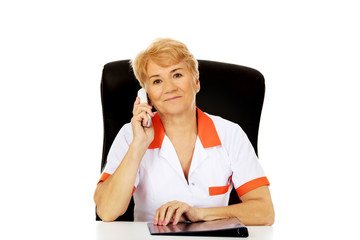 Smile elderly female doctor or nurse sitting behind the desk and talking through a phone